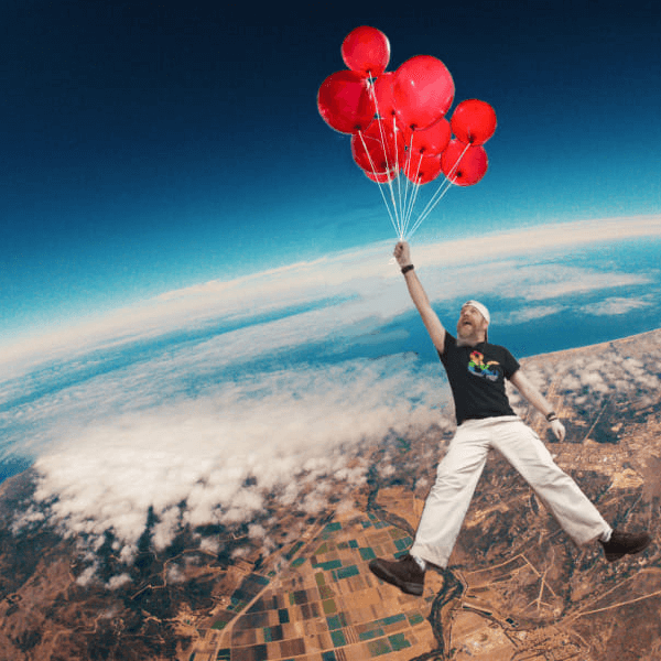 meet our staff, Stevil SteviL of Noble Knight Games is shown as a tall man in a Dungeons & Dragons logo tee, holding a bunch of red balloons by the string, while he floats happily miles above the earth