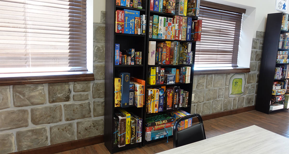 a bookshelf full of tabletop games up against a wall that is painted to look like stone
